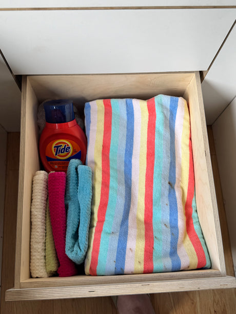 Compact RV rental storage: an open drawer holds Tide detergent, various cleaning cloths, and a striped towel.