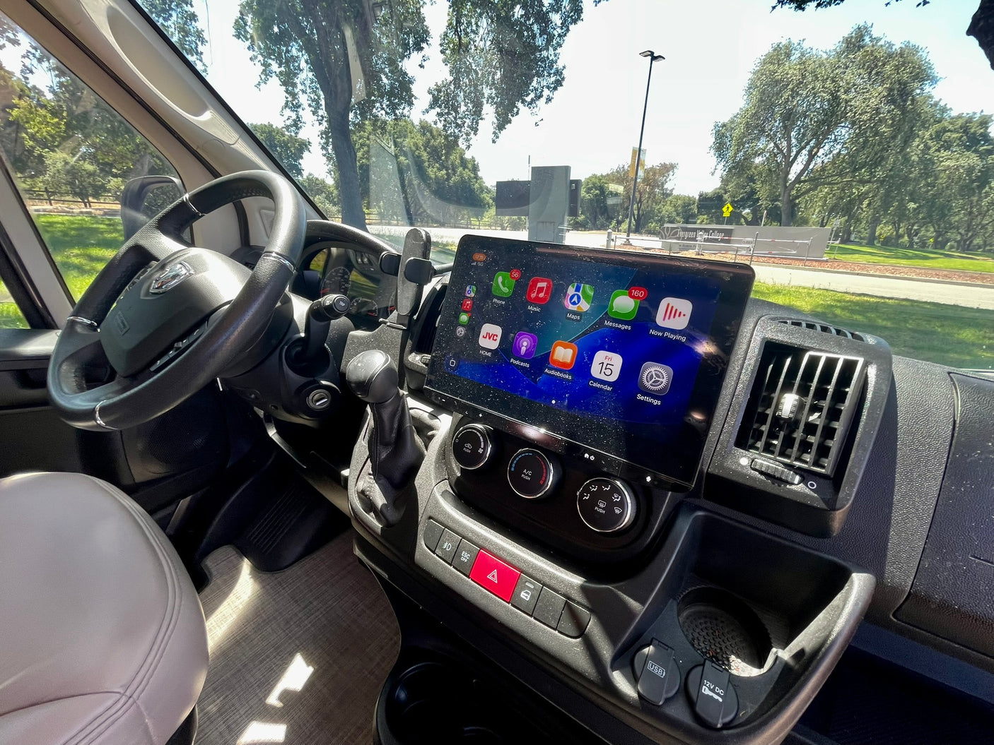 Inside an RV rental: dashboard with a large JVC screen, Apple CarPlay apps for navigation/music, steering wheel, and trees outside.