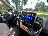 Inside an RV rental: dashboard with a large JVC screen, Apple CarPlay apps for navigation/music, steering wheel, and trees outside.