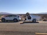 White SUV towing an angular, modern camper trailer. Parked roadside in a vast desert; consider this RV rental for trips.
