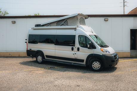 Silver camper van with pop-top roof, ideal for RV rental, parked on asphalt by a white building.