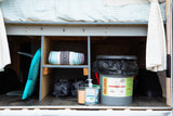 Organized storage in an RV rental, featuring a surfboard, blanket, portable toilet, and hand sanitizer for on-the-go convenience.