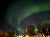 Vibrant green Northern Lights streak across a dark night sky over trees & buildings. An RV rental is perfect for aurora viewing!