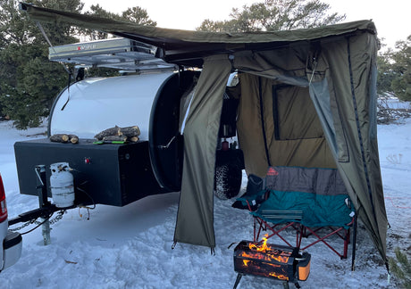 Cozy winter camping with an RV rental teardrop trailer, extended awning, and warm fire pit in the snow.