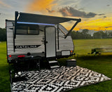 RV rental: Catalina travel trailer with lit awning, outdoor rug, grill, and cooler on a grassy field at sunset. Perfect for camping.