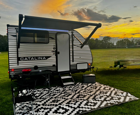 RV rental: Catalina travel trailer with lit awning, outdoor rug, grill, and cooler on a grassy field at sunset. Perfect for camping.