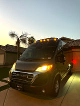 Dark grey Ram ProMaster RV rental van in a driveway at dusk. Headlights and amber roof lights are illuminated.