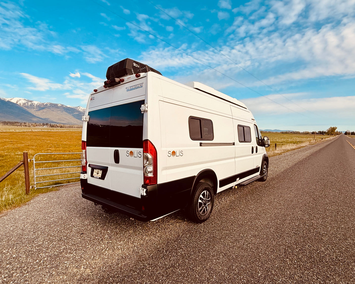 This white Solis rv rental van offers adventure. Parked roadside with mountains & fields, ready for camping & scenic drives.