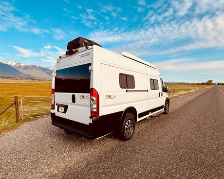 This white Solis rv rental van offers adventure. Parked roadside with mountains & fields, ready for camping & scenic drives.
