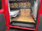 Red RV rental interior with a cozy bed, compact kitchen, wood floor, and tropical curtains.