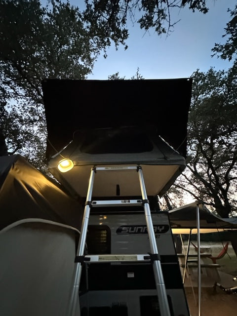 RV rental travel trailer with rooftop tent, illuminated by an outdoor light, and ladder for easy camping access.