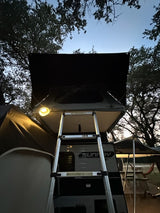 RV rental travel trailer with rooftop tent, illuminated by an outdoor light, and ladder for easy camping access.