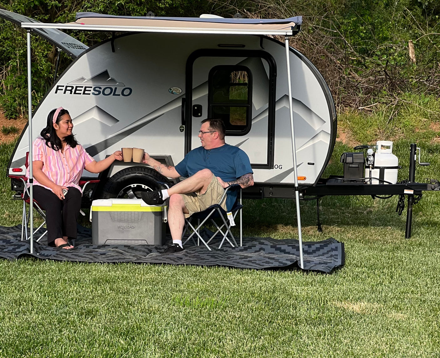 A couple relaxes by their Freesolo RV rental, a teardrop camper under an awning with chairs and a mat, enjoying outdoor leisure.