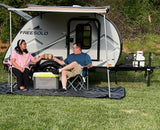 A couple relaxes by their Freesolo RV rental, a teardrop camper under an awning with chairs and a mat, enjoying outdoor leisure.
