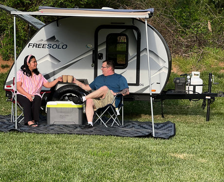 A couple relaxes by their Freesolo RV rental, a teardrop camper under an awning with chairs and a mat, enjoying outdoor leisure.