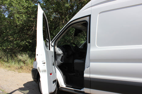 This white RV rental van has its driver's door open, revealing the interior cabin, parked on a gravel path near green trees.