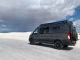 Explore White Sands with an RV rental! Dark custom camper van on white road, flanked by gypsum dunes under dramatic sky.