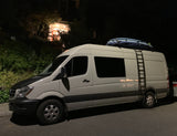 A white RV rental van, topped with surfboards and a ladder, parked on a residential street at night.