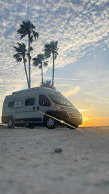 An RV rental camper van parked on gravel, silhouetted by palm trees and a vibrant sunset under a textured sky.