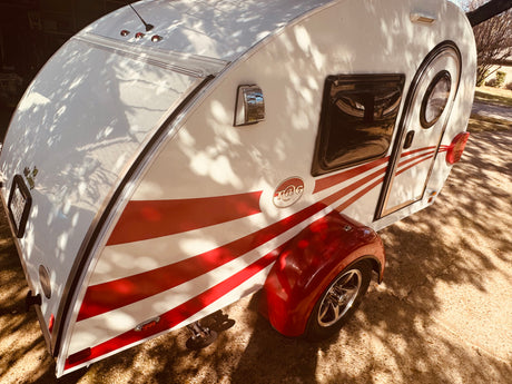 White T@G teardrop camper with red stripes, ready for an RV rental journey. Parked outside with tree shadows.