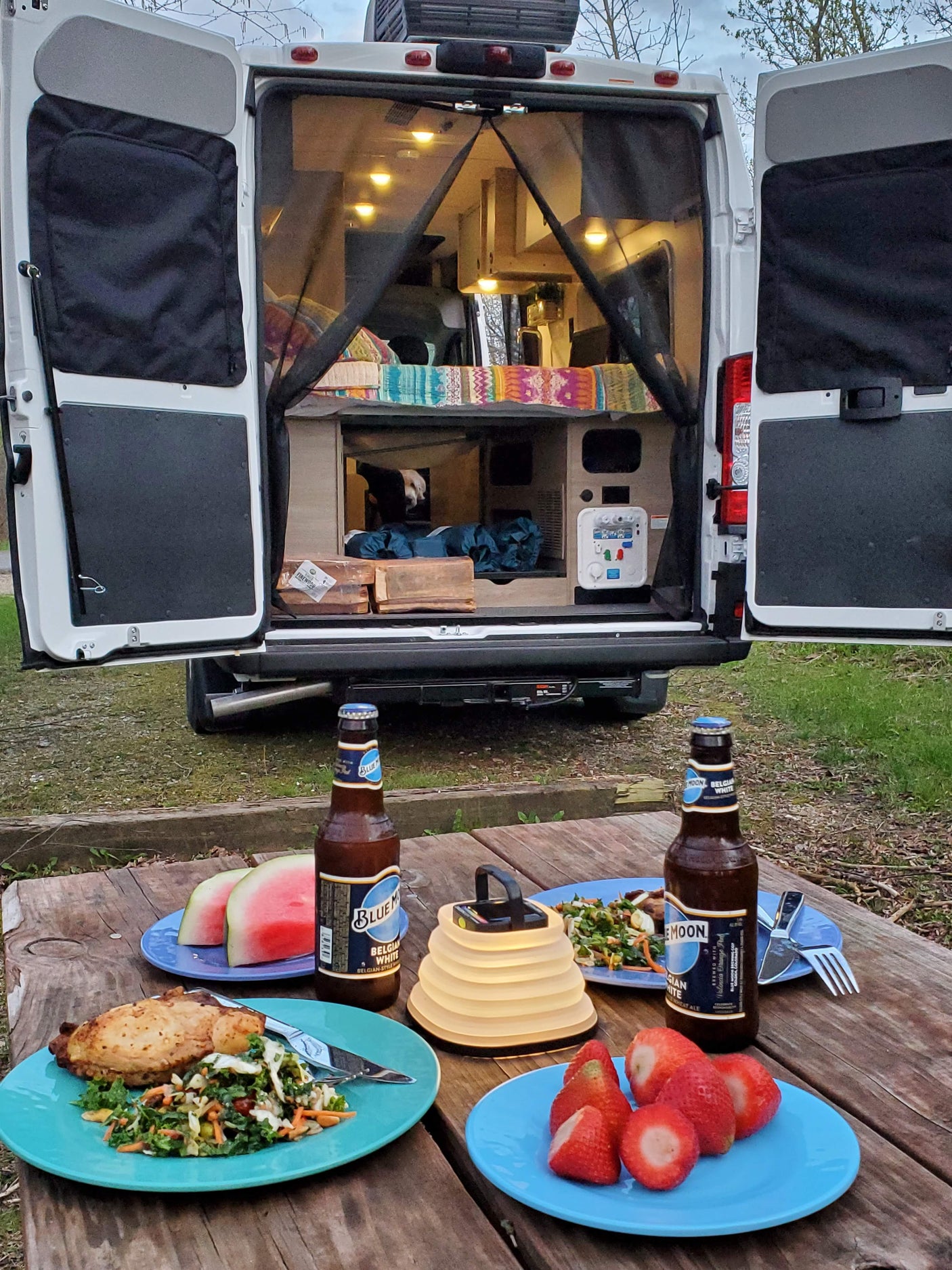 Enjoying a campsite dinner from an RV rental: Open van doors, a picnic table with food, beer, fruit, and a lantern.