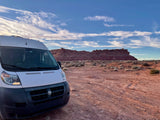 A white camper van (RV rental) on a red dirt road, facing a large red rock mesa under a blue sky with wispy clouds.