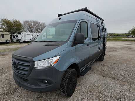 RV rental: A grey Mercedes-Benz Sprinter camper van with a black hood & off-road tires, parked on a gravel lot with other RVs.