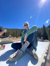 Smiling woman wearing sunglasses and a child on an RV rental roof, with bright blue sky and pine trees.