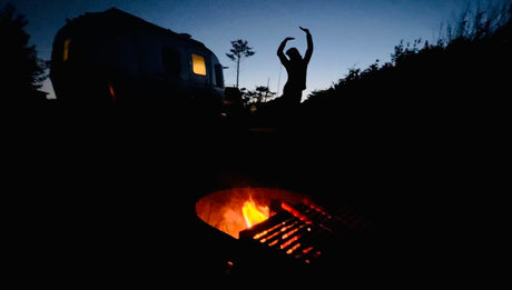 Cozy RV rental camping: A glowing campfire, silhouetted figure, and illuminated trailer under the night sky.