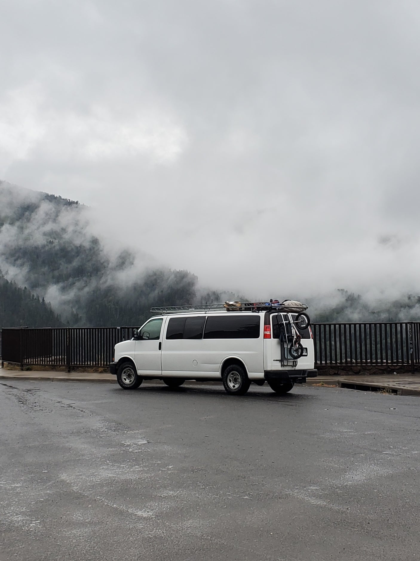 White camper van (RV rental) with gear parked on wet asphalt, overlooking misty, tree-covered mountains on a cloudy day.