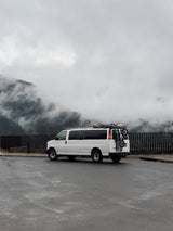 White camper van (RV rental) with gear parked on wet asphalt, overlooking misty, tree-covered mountains on a cloudy day.
