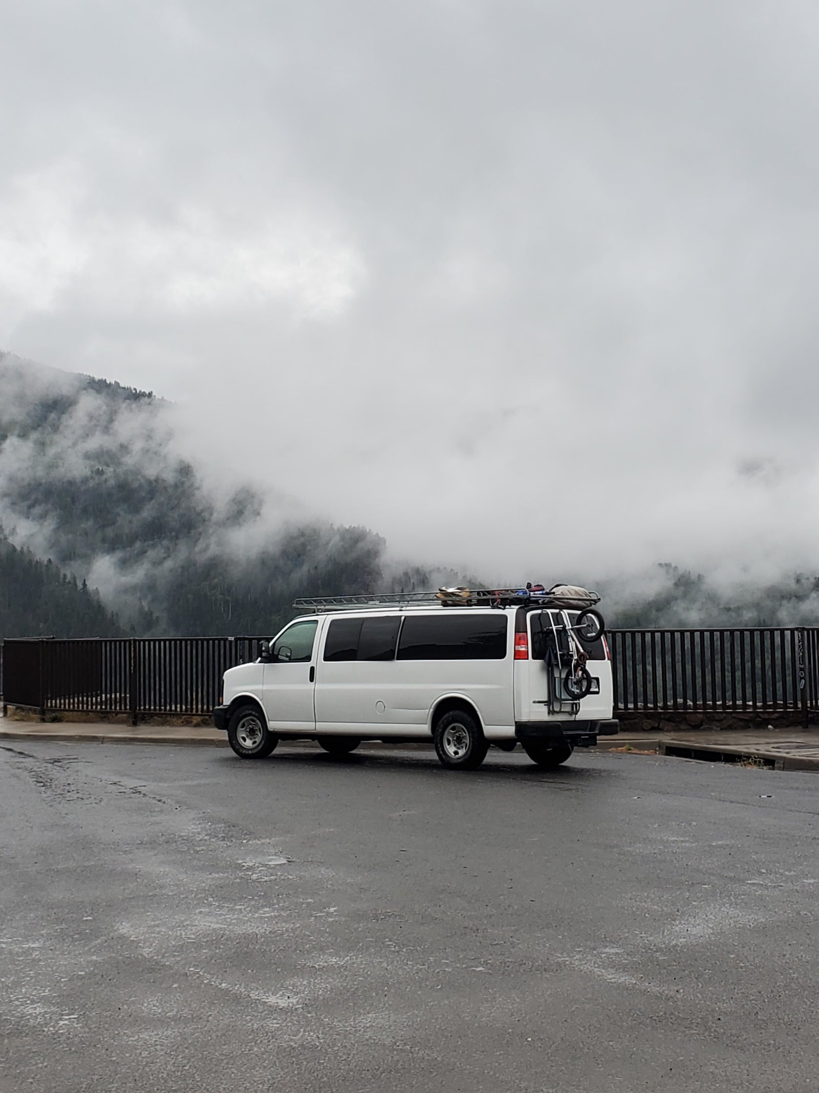 White camper van (RV rental) with gear parked on wet asphalt, overlooking misty, tree-covered mountains on a cloudy day.