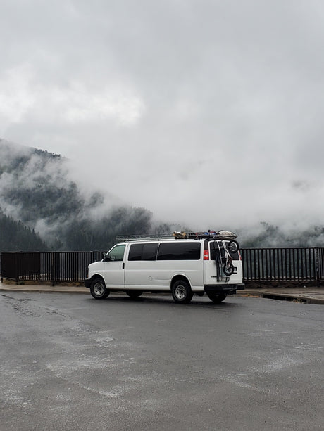 White camper van (RV rental) with gear parked on wet asphalt, overlooking misty, tree-covered mountains on a cloudy day.