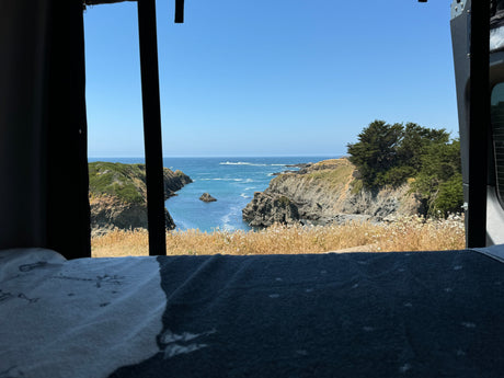 Inside an RV rental, a stunning ocean view frames rocky cliffs, blue waters, and sky, as seen from a comfortable bed.