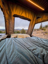 Inside a comfy RV rental rooftop tent, showcasing Klymit pillows, a cozy bed, and stunning mountain views under a cloudy sky.