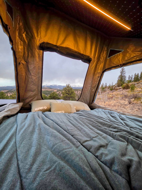 Inside a comfy RV rental rooftop tent, showcasing Klymit pillows, a cozy bed, and stunning mountain views under a cloudy sky.