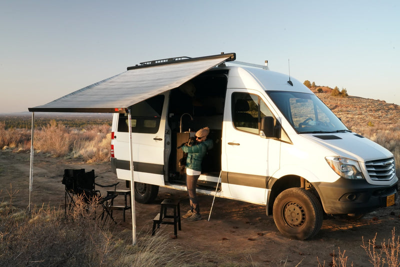 An RV rental camper van with an awning extended is parked in nature. A person holds a dog by the open side door.|Cozy RV rental interior with a comfortable bed — photo 1