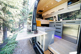 This spacious rv rental camper van interior features a kitchen, bed, and outdoor table, parked amidst a serene forest.