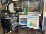 An RV rental's pull-out outdoor kitchen features a stove cooking, sink, and open fridge with food, plus storage.