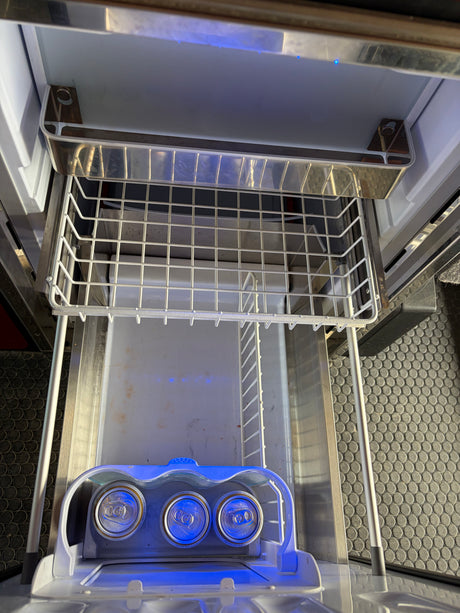 Top-down view inside an RV rental refrigerator with three canned beverages, an empty ice maker tray, and blue LED lights.