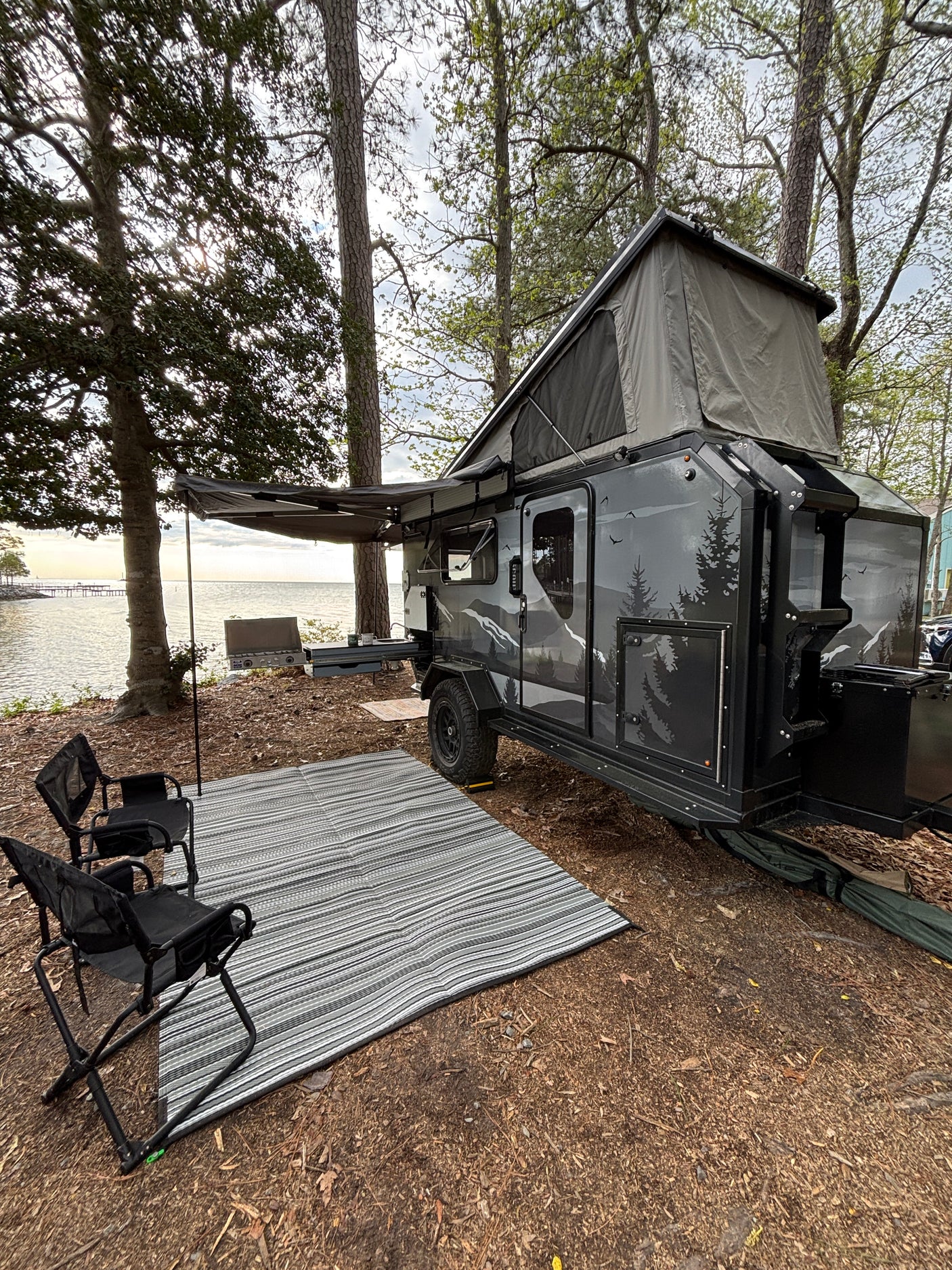 This RV rental features a pop-up camper trailer by the water with an awning, outdoor mat, and camp kitchen setup.