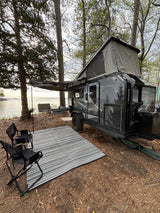 This RV rental features a pop-up camper trailer by the water with an awning, outdoor mat, and camp kitchen setup.
