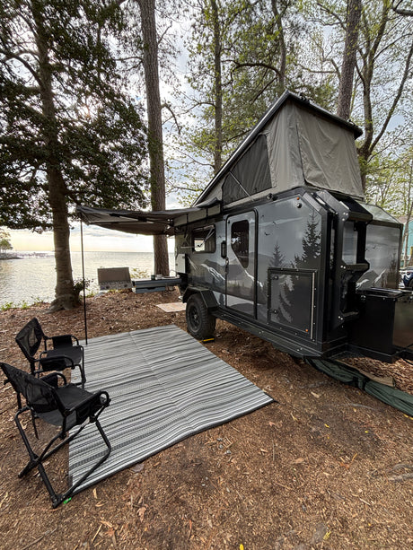 This RV rental features a pop-up camper trailer by the water with an awning, outdoor mat, and camp kitchen setup.