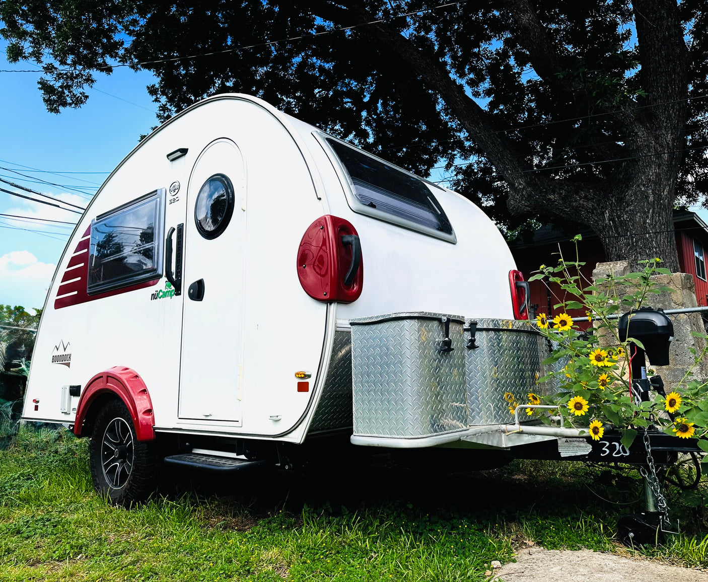 White teardrop RV rental with red accents, parked under a large tree with sunflowers. Perfect for outdoor adventures!