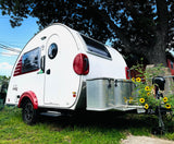 White teardrop RV rental with red accents, parked under a large tree with sunflowers. Perfect for outdoor adventures!