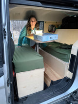 A smiling woman showcases the versatile interior of her RV rental, featuring beds, a table, and a board game.
