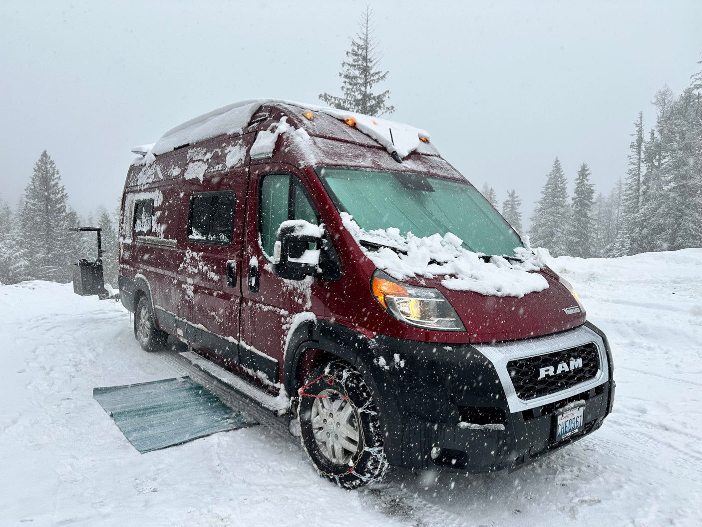 Maroon RV rental van, covered in snow with tire chains and glowing lights, parked among snowy pine trees during a snowstorm.