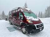 Maroon RV rental van, covered in snow with tire chains and glowing lights, parked among snowy pine trees during a snowstorm.