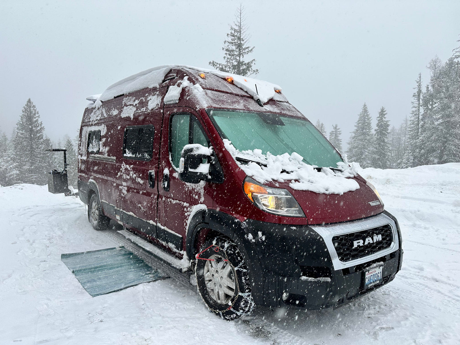 Maroon RV rental van, covered in snow with tire chains and glowing lights, parked among snowy pine trees during a snowstorm.