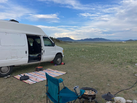 Relaxing campsite with an rv rental camper van, a cozy fire pit, and beautiful mountain views under a blue sky.
